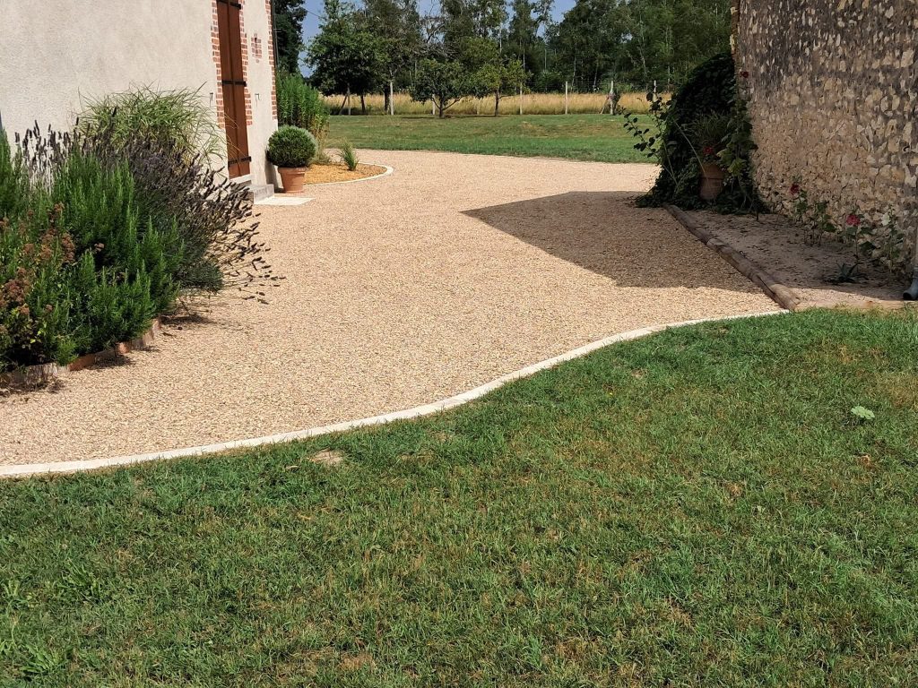 terrasse gravillonnée beige avec plantes en bordure d'une maison. Pelouse verte tondue au premier plan et en fond.