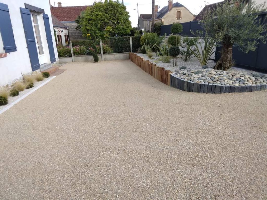 cour gravillonnée beige avec bordure plantée au fond et habitation sur la gauche