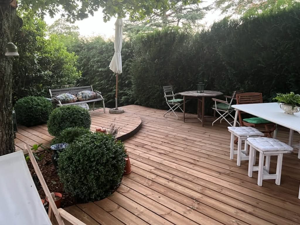 Terrasse en bois aménagée avec buissons, haie dans le fond, table et chaises blanches et chaise longue devant
