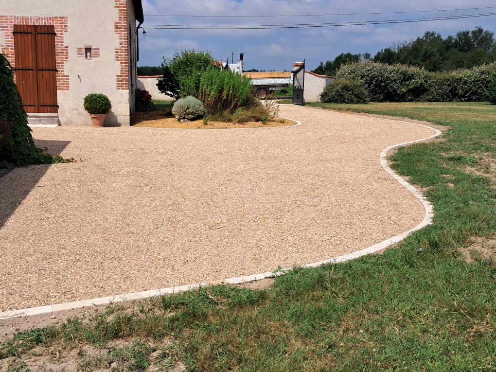 Cour et allée en gravillons beige avec pelouse au premier plan et habitation dans le fond.