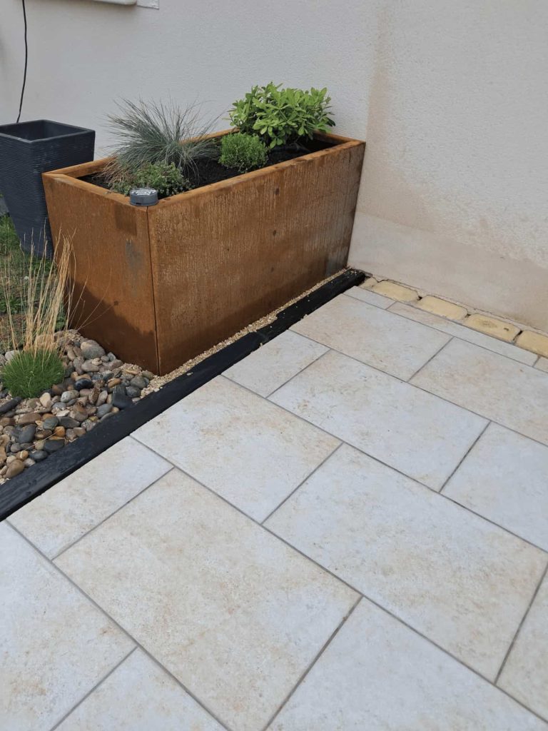 Coin de terrasse carrelée en blanc avec jardinière métallique brune sur le côté.